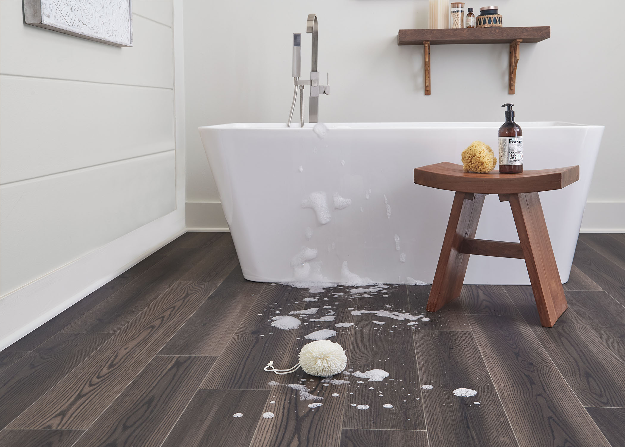 dark brown waterproof rigid vinyl plank floor in bathroom with white rectangle bathtub and dark wood stool plus soapy water running down front of tub and soapy scrub sponge on floor