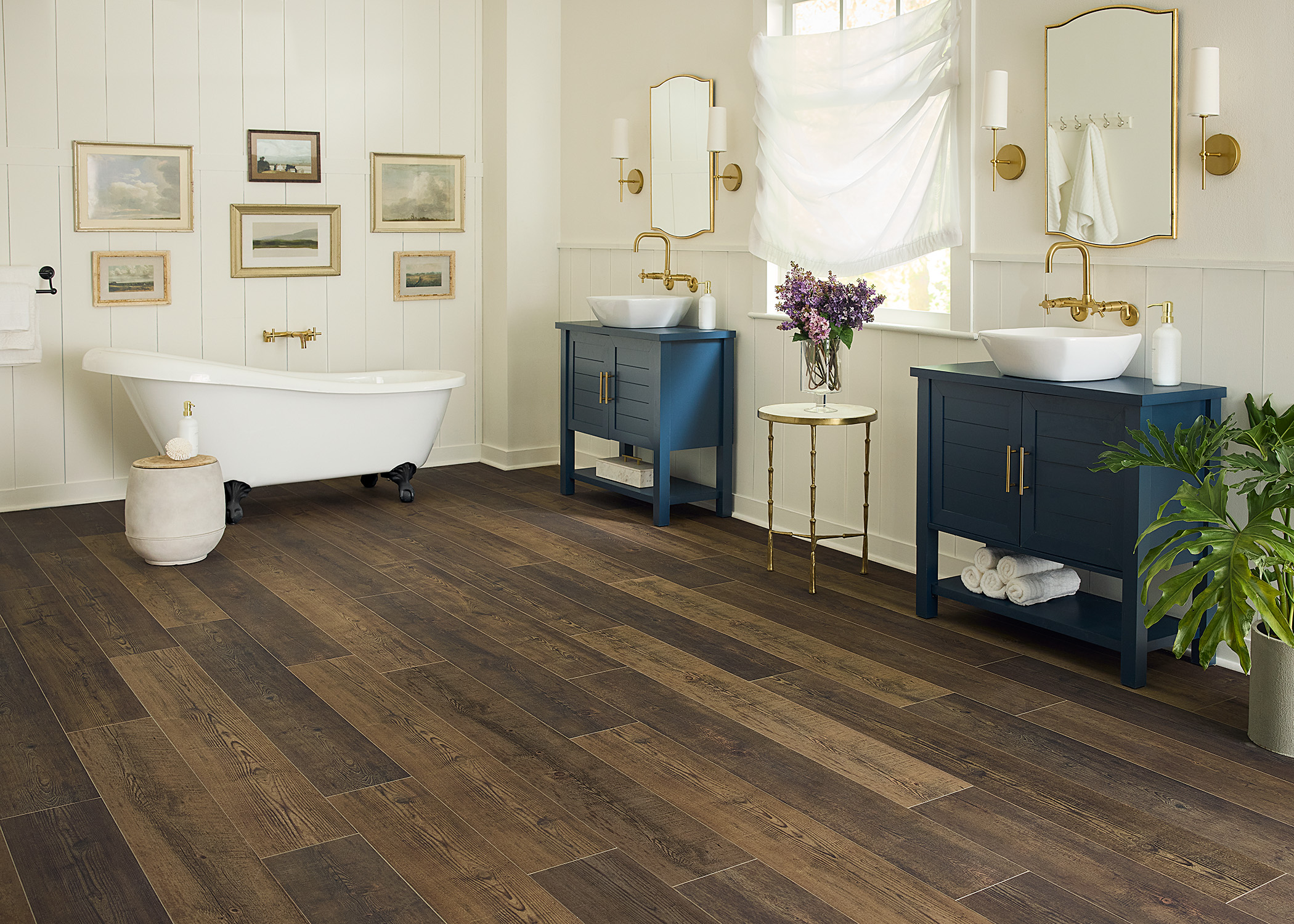 multi toned brown waterproof rigid vinyl plank floor in bathroom with matching dark blue vanities with vessel sinks plus freestanding clawfoot tub with antique artwork on shiplap wall above and sheer balloon curtain on window