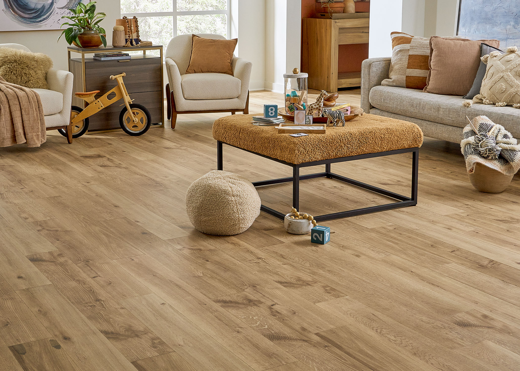 blonde waterproof rigid vinyl plank flooring in living room with beige furnishings plus gold upholstered ottoman and wooden tricycle and knotting material in a basket