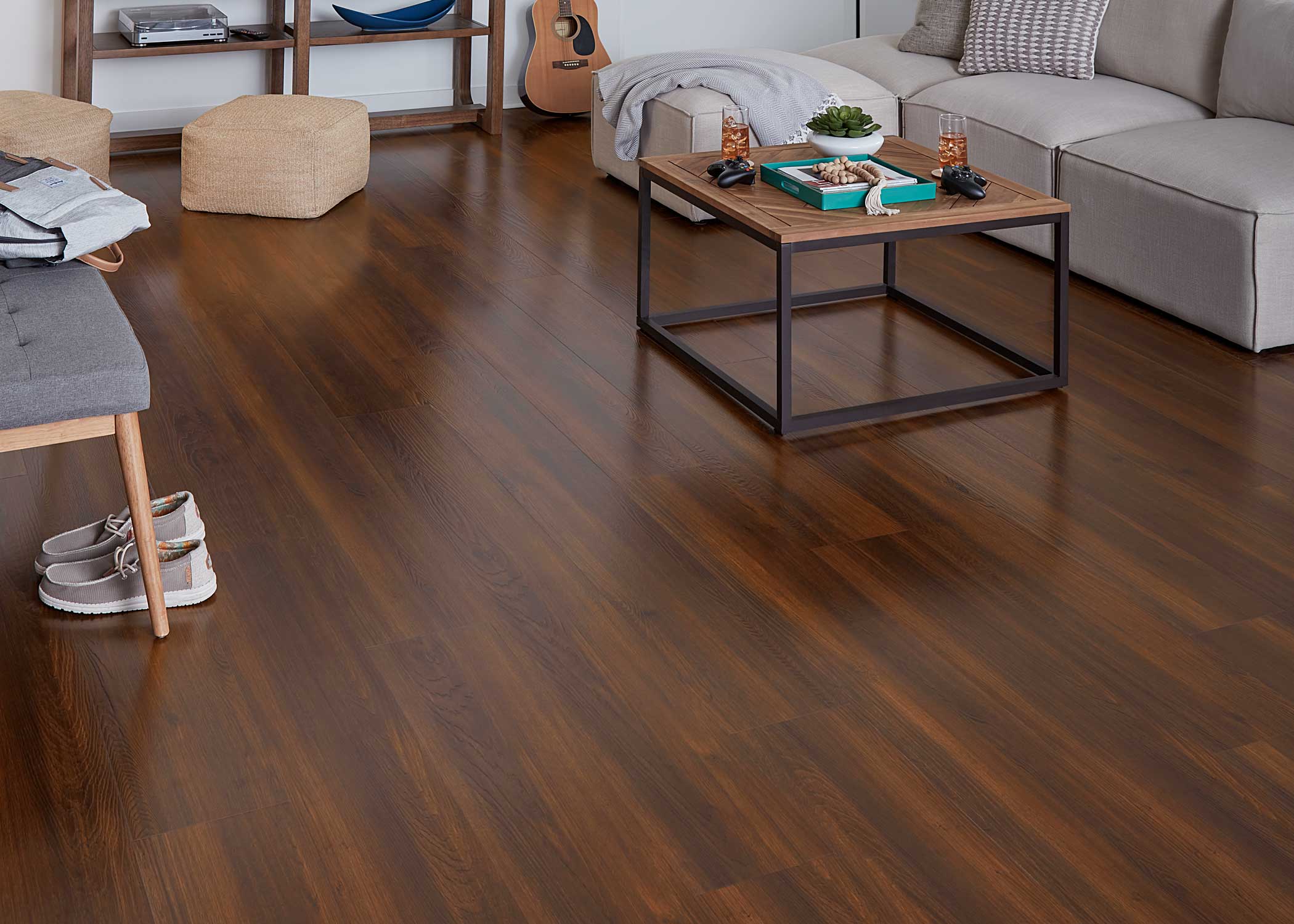 bronze waterproof rigid vinyl plank flooring in lower level family room with beige armless sectional with wood coffee table plus small bench with upholstered seat and dark wood console with two tan square ottomans in front