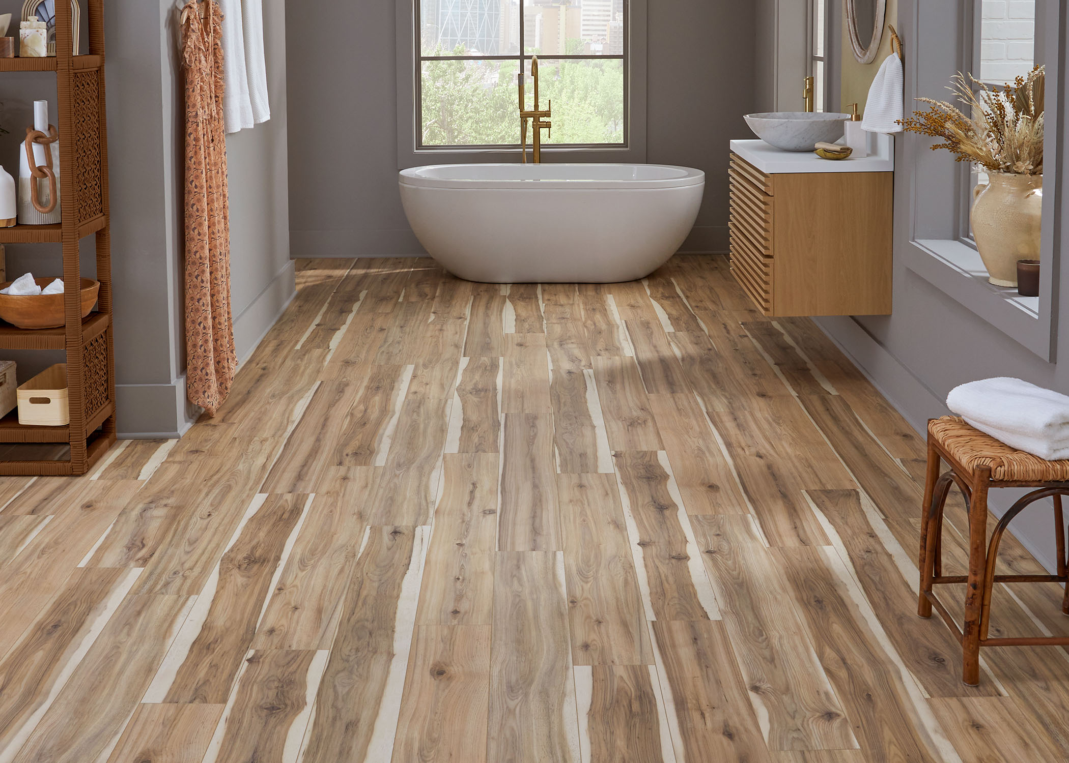 multi toned brown waterproof rigid vinyl plank flooring in bathroom with blonde wood floating vanity plus freestanding oval tub and rattan and cane stool