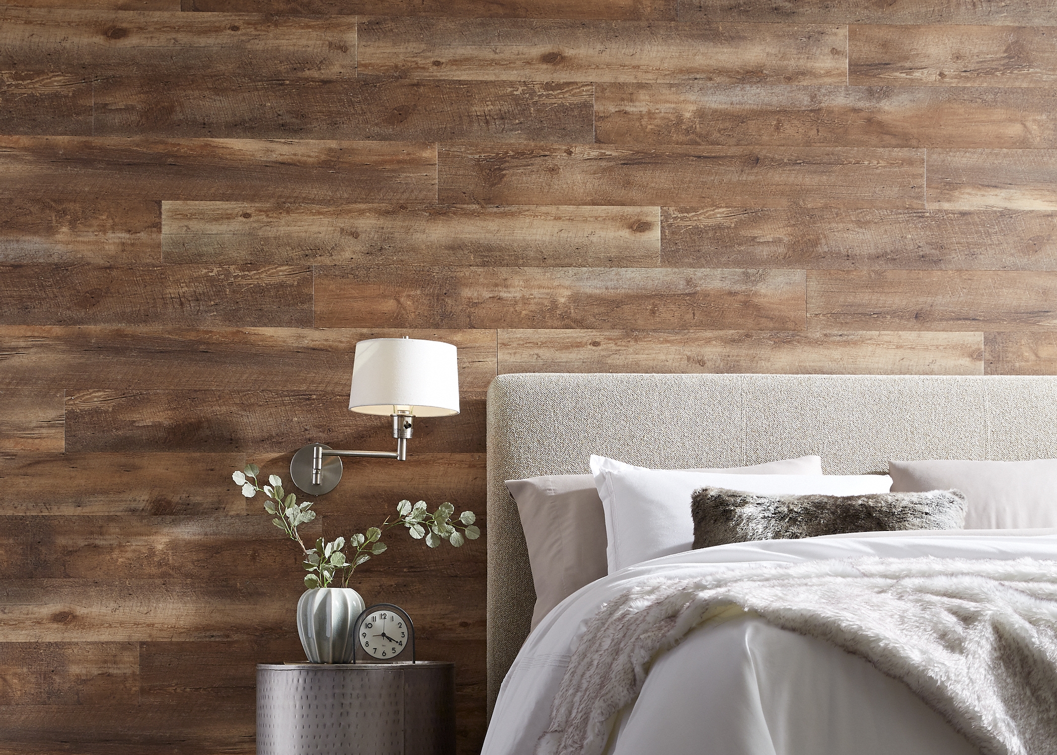 multi tone brown waterproof luxury vinyl plank flooring on wall in bedroom behind bed with beige upholstered headboard and cream bedding plus silver side table and faux fur accent pillow