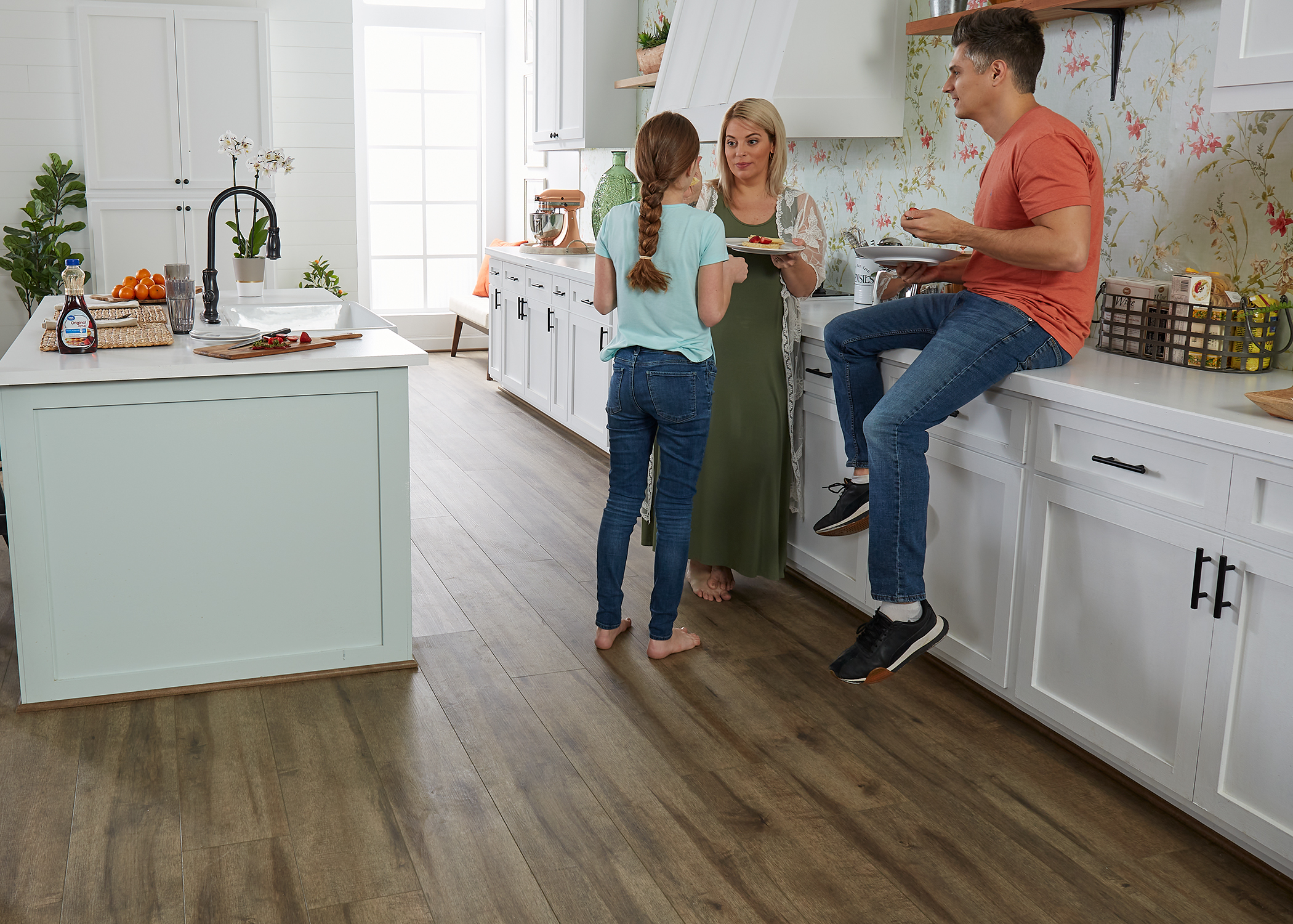 multi tone brown waterproof rigid vinyl plank flooring in kitchen with white cabinets and countertops plus floral wallpaper and family enjoying breakfast next to oversized island