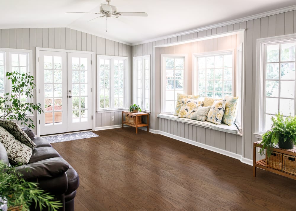 3/8 in. x 4.75 in. Abilene Hickory Quick Click Engineered Hardwood Flooring in sunroom with built in window seat with blue and yellow flower pillows plus light gray painted shiplap and brown leather sofa