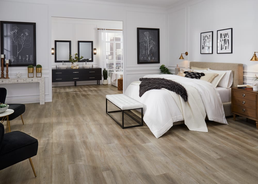 9mm with Pad Chapel Bridge Oak Waterproof Hybrid Resilient Flooring in primary bedroom with tan upholstered headboard plus white bedding and black and white artwork plus view into bathroom with black dual vanity