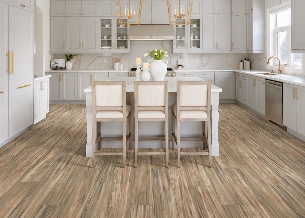 7mm with Pad Summer Plains Acacia Waterproof Rigid Vinyl Plank Flooring in kitchen with light gray cabinets plus oversized island with three chairs and white vase with flowers on counter