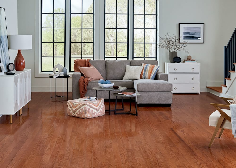 3/4 in x 3.25 in Gunstock Oak Solid Hardwood Flooring in lower level family room with taupe sectional with colorful accent pillows plus orange square ottoman and white credenza