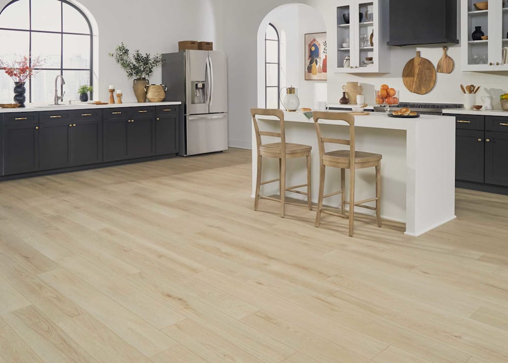 8mm with Pad Goldfinch Oak Waterproof Hybrid Resilient Flooring in kitchen with dark gray cabinets with white countertops plus white island and blonde wood barstools