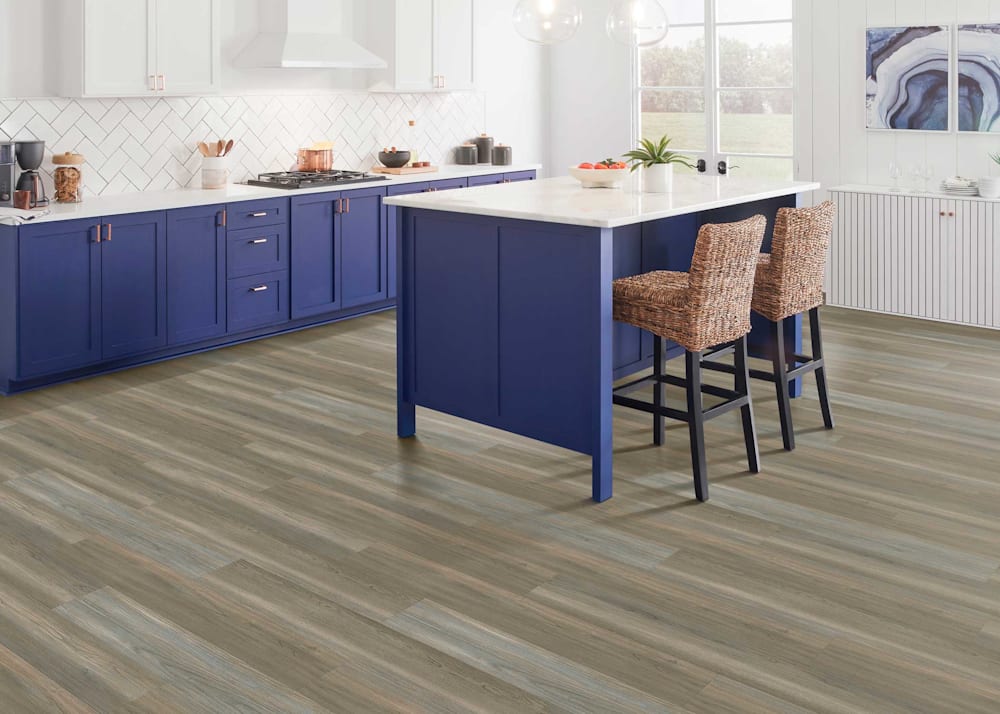 5mm Eastlake Oak Waterproof Luxury Vinyl Plank Flooring in kitchen with blue lower cabinets with white countertops and white upper cabinets plus blue island with white countertop and brown rattan bar stools